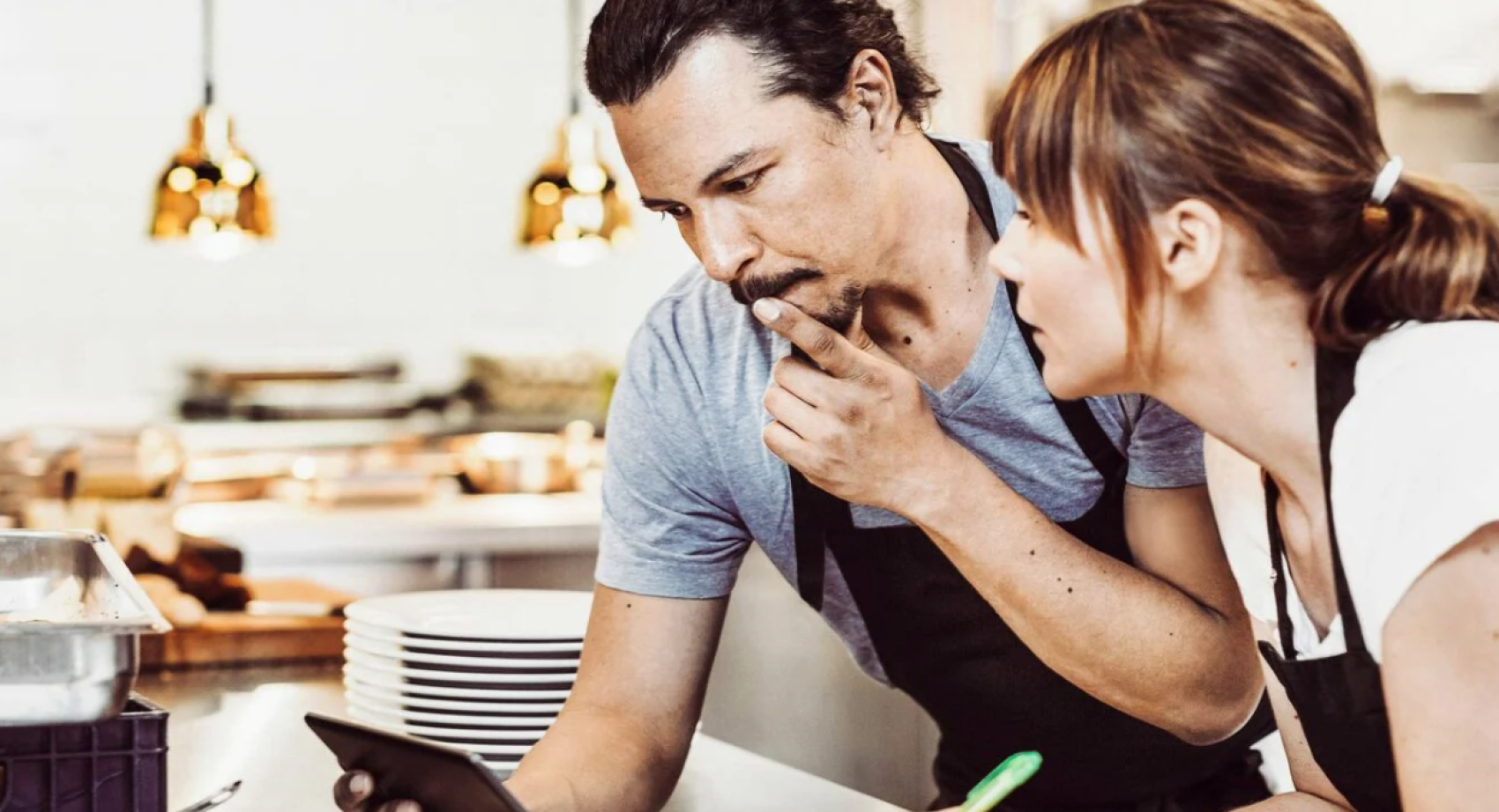 Deux restaurateurs en tablier se tiennent à un comptoir et regardent attentivement un smartphone. L'un d'eux a une expression pensive, se touchant le menton, tandis que l'autre se penche pour voir l'écran. Des assiettes et des ustensiles de cuisine sont à l'arrière-plan.