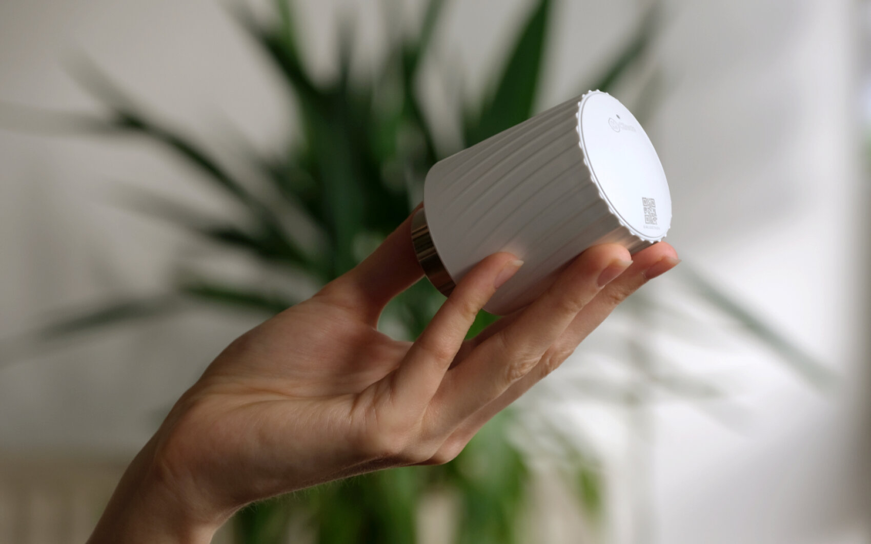 Eine Hand hält ein kleines, weißes, zylindrisches elektronisches Gerät mit einer gerippten Textur vor einem unscharfen Hintergrund mit grünen Pflanzenblättern.