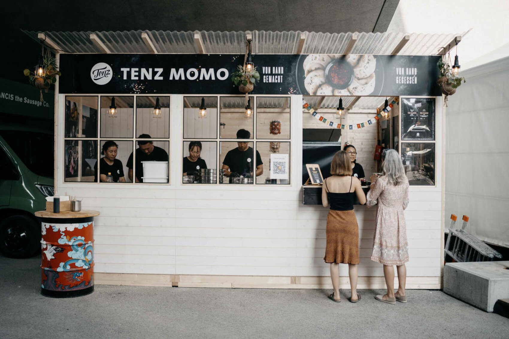 Un stand de nourriture blanc nommé Tenz Momo avec cinq employés à l'intérieur, décoré de lumières et de drapeaux. Deux femmes se tiennent au comptoir pour commander de la nourriture. Un tonneau coloré se trouve devant l'étal.
