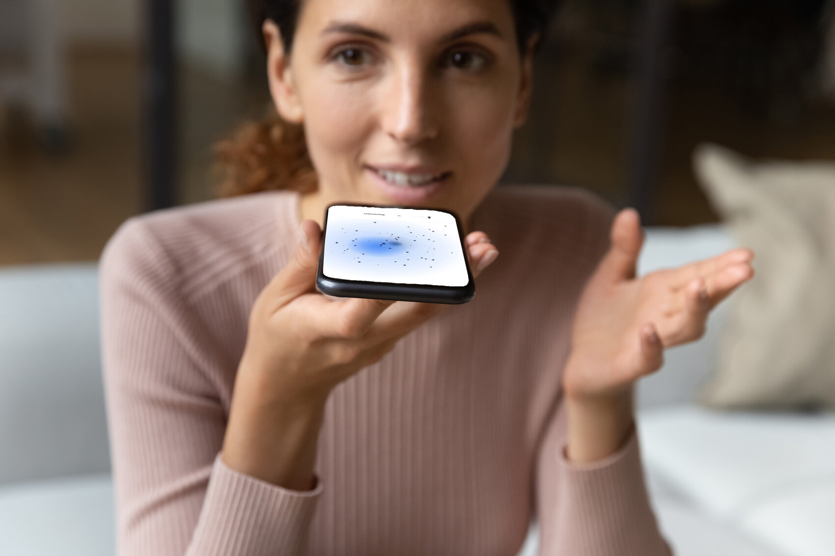 Eine Frau sitzt in einem Haus, hält ein Smartphone waagerecht und spricht in das Gerät. Der Bildschirm des Telefons zeigt eine Sprachassistenten-Schnittstelle mit einem leuchtenden Licht und Punkten an. Sie hat einen ausdrucksstarken, engagierten Blick.